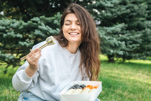 Cheerful young woman eating sushi in the park, picnic in nature, sushi delivery concept. - Valokuva, kuva