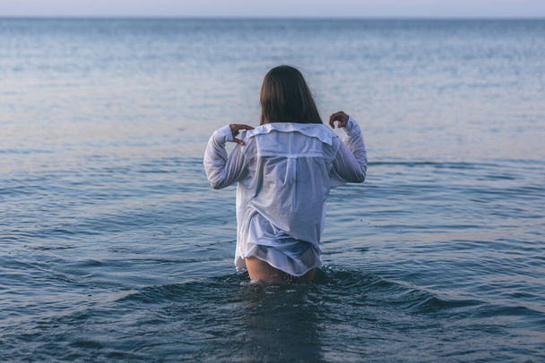 A woman in a swimsuit and a white shirt in the sea at sunset, view from the back. - Foto, afbeelding