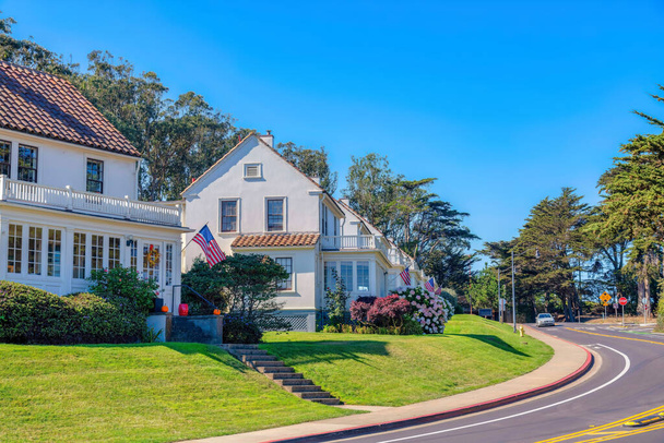 Suburban neighborhood with sloped lawn near the curved street in San Francisco, CA. Home facades with USA flags and ornamental shrubs displayed at the front near the road with signs and car. - Zdjęcie, obraz