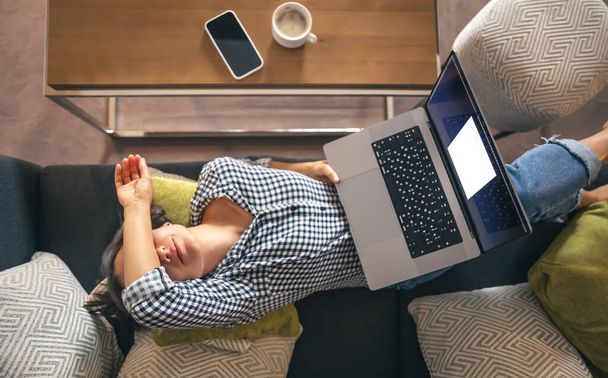 Κουρασμένος από την εργασία, μια νεαρή γυναίκα με ένα φορητό υπολογιστή βρίσκεται στον καναπέ, θέα από ψηλά. - Φωτογραφία, εικόνα