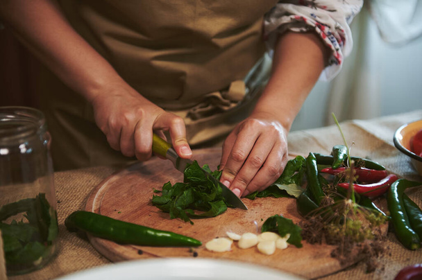 Close-up of the hands of an unrecognizable woman, housewife in a beige apron, chopping fresh culinary herbs on a wooden cutting board while preparing brine for pickling chili peppers at home kitchen - Foto, Imagen