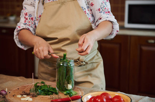 Close-up of a woman, housewife wearing a beige chefs apron, filling the glass jar with fresh chili peppers, cooking marinated food, making preserved delicacies for the winter season at home kitchen - Foto, imagen