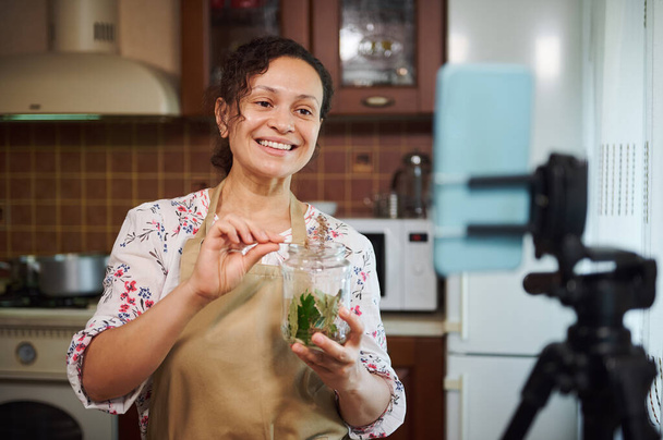 Pleasant woman, housewife in a chefs apron, looks at a smartphone camera, mounted on a tripod and records video content for her food blog. Canning. Pickling. Marinating. Making homemade pickles - Foto, afbeelding