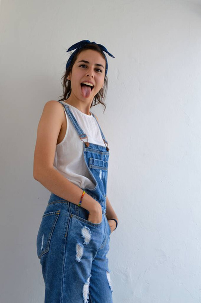 A vertical shot of a self-confident girl in blue overalls being silly and sticking her tongue out at the camera - 写真・画像