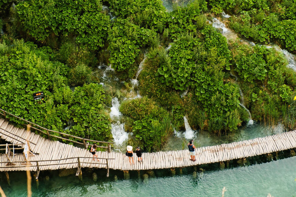 Wooden footbridge between aquamarine lake and hilly green bank. Tourists discover nature of famous national natural park reserve on Plitvice lakes upper view - Foto, Imagem