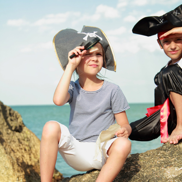 Sad boy and girl in a pirate costume and a beautiful pirate hats sitting on a large rock at the seaside - Foto, Imagen