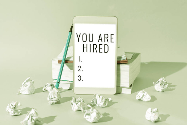 Sign displaying You Are Hired, Business overview Recruitment being selected for a job the right candidate Important Messages Presented On Mobile Phone Leaning On Books And Notebook. - Photo, Image