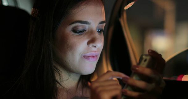 Young woman at night in the back seat of a car checking smartphone typing - Photo, Image