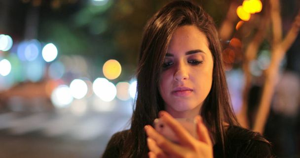 Young hispanic brunette woman checking her cellphone at night. Latina girl holding her smartphone in the evening next to traffic lights - Φωτογραφία, εικόνα