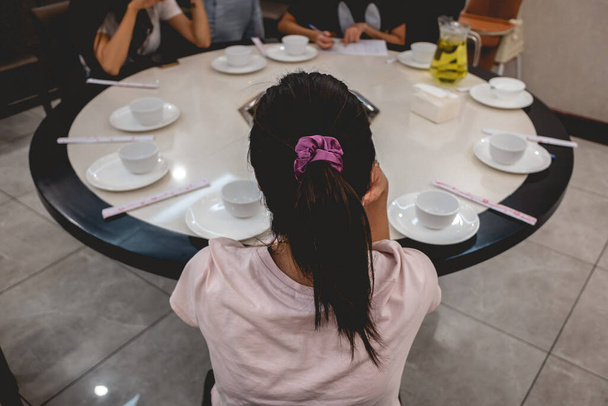 Beautiful young asian girl with ponytail and satin scrunchie hair tie seated in front of a chinese round table with empty white plates and group of people - Fotó, kép