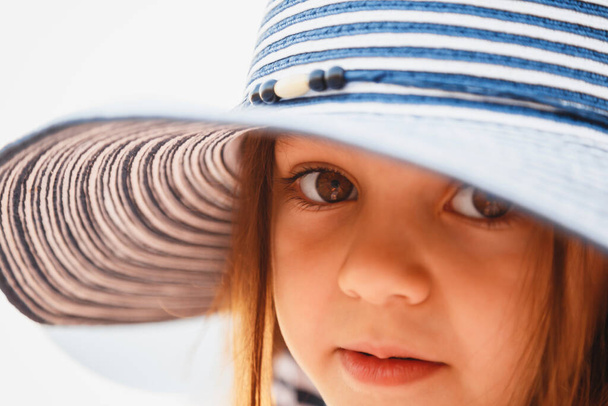 Close up portrait of cute little girl with big brown eyes in striped sunhat looking at camera. Selective focus. - Foto, Bild