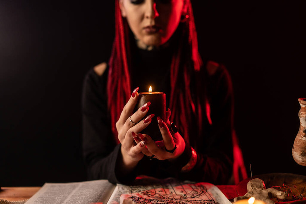 A witch who performs a ritual with a candle. On the table is a spell book and a voodoo doll. A witch who practices shamanism. - Zdjęcie, obraz