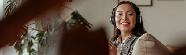 Ελκυστική Ασιάτισσα επιχειρηματίας που ακούει μουσική καθισμένη με φορητό υπολογιστή στο γραφείο και ψάχνει κάμερα - Φωτογραφία, εικόνα