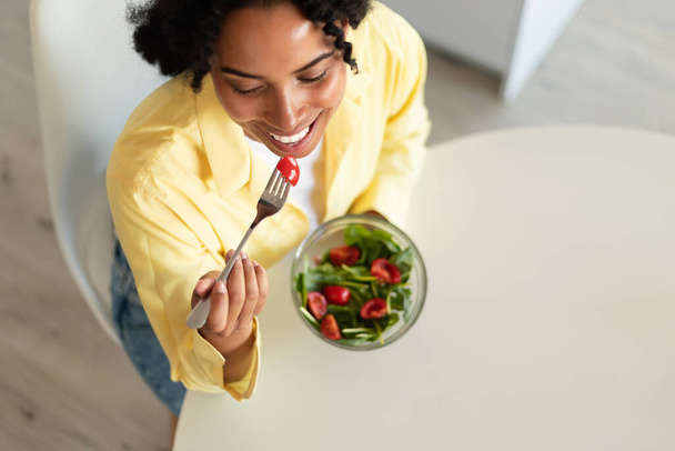 Fit african american lady eating salad with organic vegetables in light kitchen interior, top view, free space. Cooking proper nutrition for health care at home - Photo, image