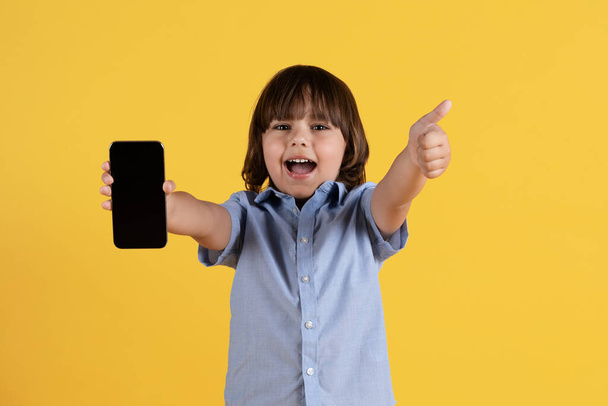 Υπέροχο παιδικό περιεχόμενο. Studio πορτρέτο του ενθουσιασμένοι μικρό αγόρι δείχνει smartphone με κενή οθόνη στην κάμερα και gesturing αντίχειρα επάνω, κίτρινο φόντο, mockup - Φωτογραφία, εικόνα