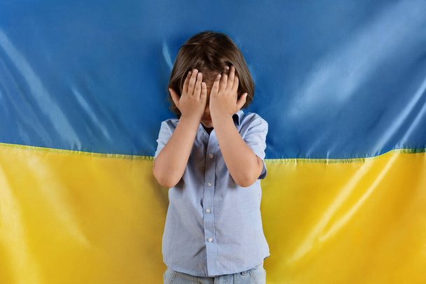 Хватит убивать детей на войне. Маленький напуганный кавказский мальчик плачет, закрывая лицо руками, позируя над украинским флагом - Фото, изображение
