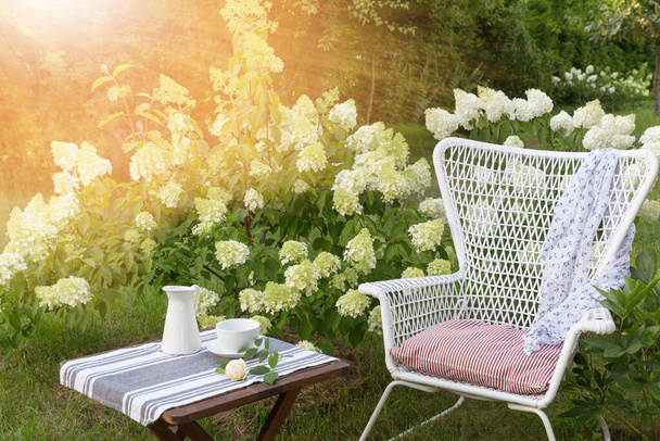 Romantic seating area in the garden, wooden table and white armchair near large flowering hydrangea bushes - Fotoğraf, Görsel