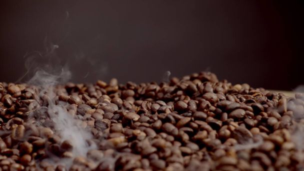 Dampf steigt in Superzeitlupe über heißen Kaffeekörnern auf. In der Nähe duftende braune Samen, die auf dunklem Hintergrund rösten. Prozess der Zubereitung aromatischer Bohnen für ein energisches Morgengetränk. Arabica-Konzept. - Foto, Bild