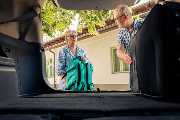 Старшие люди грузят багаж в багажник автомобиля, готовясь к отъезду на летнем отдыхе на автомобиле. Пара путешествий на отдых дорожное путешествие в пенсионном возрасте, собирается на приключение назначения. - Фото, изображение