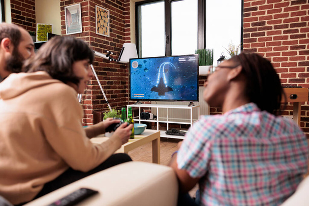 Багатоетнічна група друзів, які сидять вдома у вітальні, граючи симулятор простору відеоігор. Люди насолоджуються розважальною діяльністю на сучасному пристрої ігрової консолі, маючи пиво і закуски
. - Фото, зображення