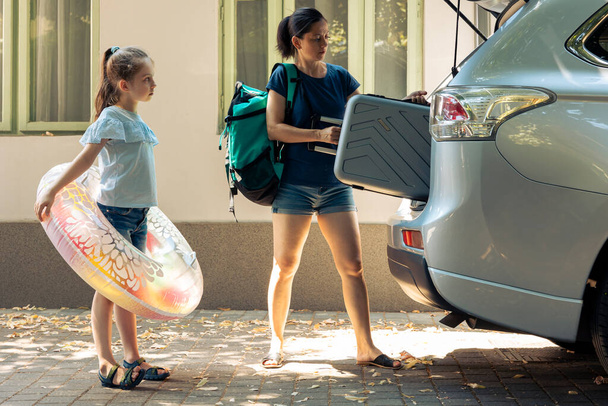 Mother and small kid travelling on vacation, loading baggage trolley and inflatable in vehicle trunk to go to seaside destination during summer holiday. Woman and little girl leaving on road trip. - Photo, image