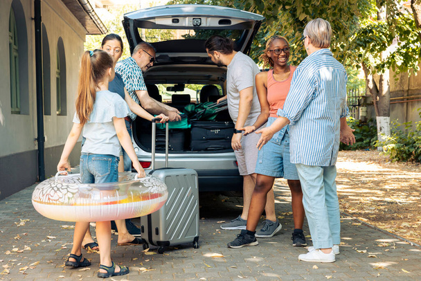 Multiethnic family and friends travelling on vacation, preparing to leave on summer holiday and loading baggage in car trunk. People sitting in driveway to go to seaside adventure trip. - Фото, зображення