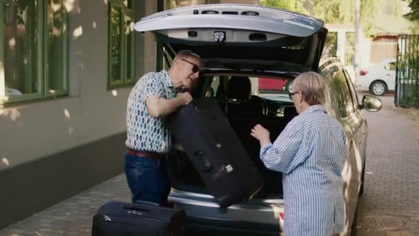 Cheerful elders going on retirement voyage while having heavy baggage and travel trolley. Senior couple putting luggage in car trunk while getting ready for holiday trip. - Video
