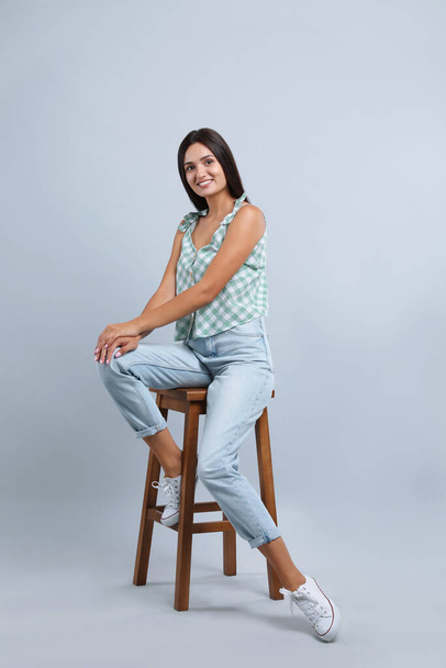 Beautiful young woman sitting on stool against light grey background - Valokuva, kuva