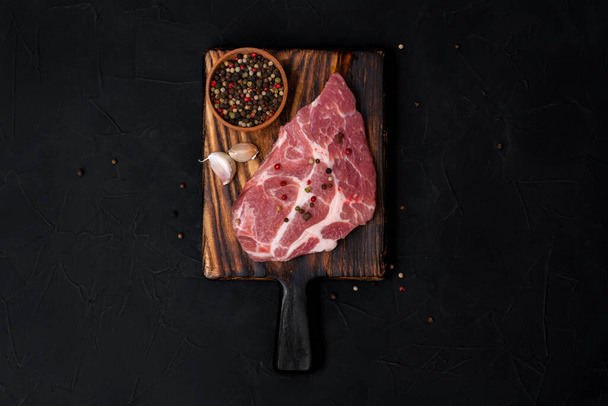 Raw pork steak on a wooden cutting board. Meat, dry pepper mixture, garlic on a black background. View from above. - Fotó, kép