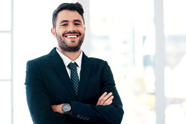 Business is all about how you play your cards. Cropped portrait of a handsome young businessman smiling while standing with his arms crossed in a modern office - 写真・画像