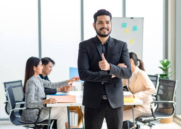 Portrait shot of millennial Indian Asian professional successful bearded businessman in formal suit standing posing smiling crossed arms in meeting room while employee and colleague brainstorming - Fotoğraf, Görsel