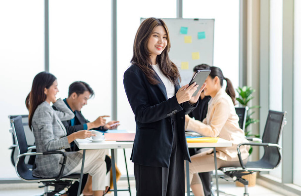 Portrait shot millennial Asian cheerful successful professional businesswoman entrepreneur in formal suit standing smiling crossed arm holding tablet computer in meeting room while employee working. - Foto, Imagen