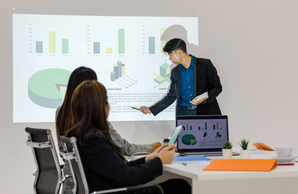 Millennial Asian young cheerful male businessman presenter in formal suit standing holding notebook and pen pointing presenting corporate graph chart data information from wall screen in meeting room. - Foto, afbeelding