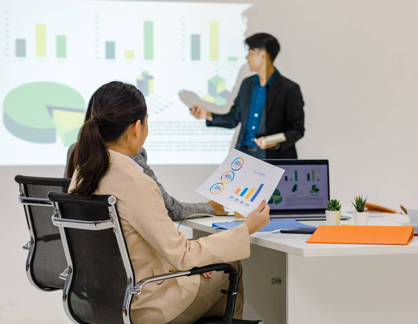 Millennial Asian young cheerful male businessman presenter in formal suit standing holding notebook and pen pointing presenting corporate graph chart data information from wall screen in meeting room. - Valokuva, kuva