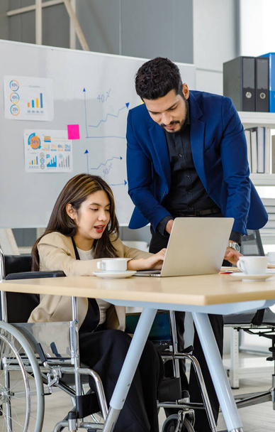 Millennial Indian male businessman manager mentor in formal suit talking helping disabled handicapped female employee sit on wheelchair working with laptop computer on table in office meeting room. - Foto, Imagen
