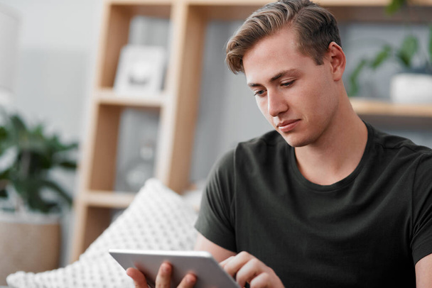 Checking what interesting events are happening around me. a handsome young man sitting alone on the sofa and using a tablet at home - Foto, imagen