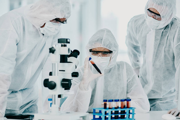 Group of scientists conducting medical research in hazmat suits and protective gloves, for coronavirus cure in a laboratory. Researchers conducting experiments and examining test tube for results - Foto, Bild