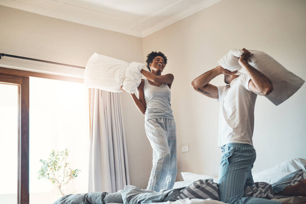 Happy, fun and playful pillow fight of a loving couple playing in their bedroom at home. Funny male and female laughing, smiling and fighting with pillows in pajamas on the bed in the morning - 写真・画像