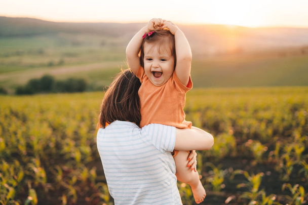 Mother holding her baby girl and playing with her among cornfield. Baby having fun. Happy family outdoors. Carefree childhood. Beautiful girl portrait. - Photo, image