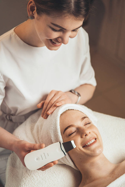 Beautician with ultrasonic scrubber cleansing womans face skin at spa salon. Electrical equipment for a spa. Apparatus for a medical patient. Acne removal. - Photo, Image
