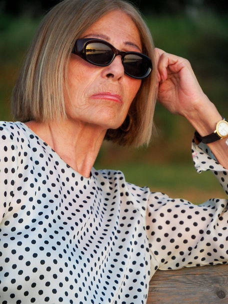 senior lady relaxing in the park wearing sunglasses near the river bank in summer - Photo, image