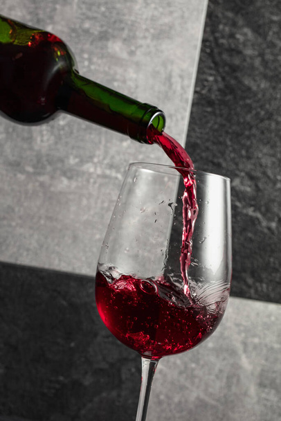 Το κόκκινο κρασί χύνεται από το μπουκάλι σε ένα ποτήρι. Γκρι και μαύρο φόντο πέτρα. - Φωτογραφία, εικόνα