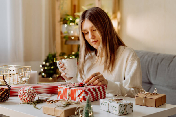 Nő csomagolás karácsonyi öko-természetes ajándék dobozok otthon. Ajándékok elkészítése fehér asztalon díszítő elemekkel, fényekkel és tárgyakkal Karácsonyi vagy újévi barkácsolás koncepció. - Fotó, kép