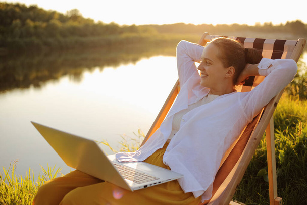 Outdoor relaxed attractive woman wearing white shirt sitting by the water in folding chair and working on laptop, sitting with hands behind head, relaxing, works during vacation. - 写真・画像