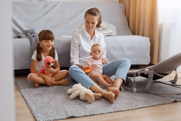 Indoor shot of cute baby, elder sister and young mother at home sitting on floor, woman wearing white shirt and jeans playing with her children during weekend. - Foto, immagini