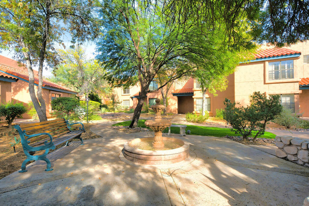 Wooden bench and fountain near the italianate houses in a subdivision at Tucson, Arizona. There are trees and lawn near the walkway leading to the entrance of the houses at the back. - Zdjęcie, obraz
