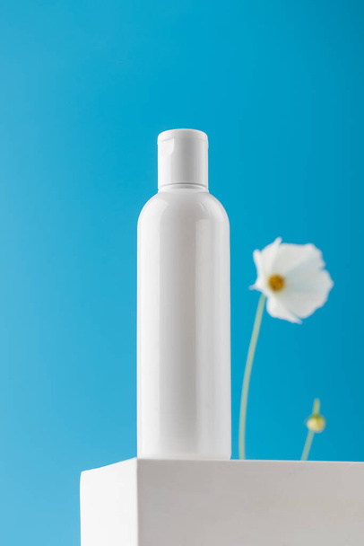 An empty bottle with a cosmetic product on a pedestal on a blue background with a delicate flower. Micellar water, facial cleansing lotion. Cleanliness and Refreshing Treatments - Photo, Image