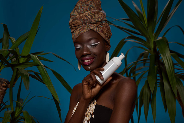 Elegant black girl holding bottle with cosmetic cream between green palms. Beautiful young slim woman wear traditional african outfit and accessories. Body and skin care. Blue background. Studio shoot - Φωτογραφία, εικόνα