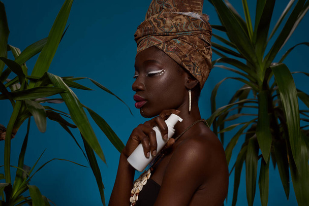 Side view of fashionable black girl with bottle of cosmetic cream between palms. Pretty young woman wear traditional african outfit and accessories. Body and skin care. Blue background. Studio shoot - Fotografie, Obrázek