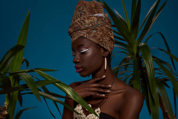Thoughtful stylish black girl posing between green palms. Attractive young slim woman wearing tank top and traditional african turban, necklace and earrings. Isolated on blue background. Studio shoot - Foto, imagen
